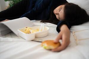 occupato e stanco donna d'affari mangiare pane per prima colazione nel letto a casa e Lavorando per consegnare finanziario dichiarazioni per un' capo. oberati di lavoro e malsano per pronto pasti, bruciato concetto. foto