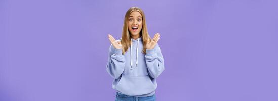 ritratto di divertito eccitato e intrattenuto contento affascinante donna con giusto capelli nel di moda felpa con cappuccio stringendo mani a partire dal diletto e sorpresa sorridente a grandi linee con gioia in posa contro viola sfondo foto