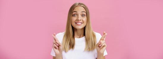 ottimista pieno di speranza eccitato ragazza con abbronzato pelle e dritto giusto capelli guardando eccitato e contento a telecamera sorridente con speranza attraversamento dita per bene fortuna fabbricazione desiderio in posa contro rosa parete foto