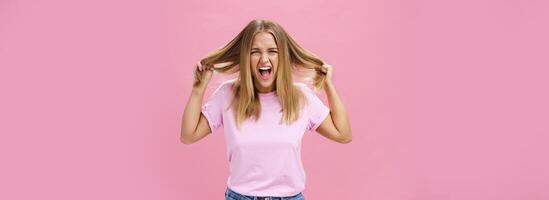 ritratto di Incazzata oltraggiato e irritata donna perdente temperare sensazione sotto pressione urlando a partire dal angoscia traino capelli su di testa in piedi infastidito al di sopra di rosa sfondo alimentato su fare taglio di capelli ogni giorno foto