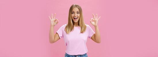ritratto di entusiasta attraente caucasico ragazza nel di moda maglietta e jeans mostrando va bene o Confermare gesto con divertito ampio Sorridi in piedi lieta al di sopra di rosa sfondo reagendo per eccellente notizia foto