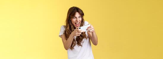 eccitato intenso giocoso gamer ragazza disadattato giocando eccezionale smartphone gioco hold mobile Telefono orizzontale smorfie desideroso vincere portico sguardo fisso schermo rubinetto Schermo In piedi giallo sfondo ribaltamento foto