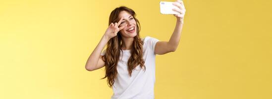 gioioso carismatico bella vista dai capelli ricci donna bianca maglietta mostrare pace vittoria cartello sorridente a grandi linee in posa tenero amichevole espressione smartphone davanti telecamera prendere autoscatto giallo sfondo foto