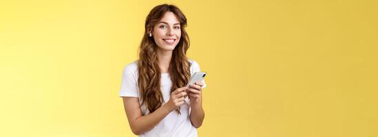 femminile bello dai capelli ricci donna bianca maglietta hold smartphone raccolto eccezionale nuovo canzone ascolta musica indossare senza fili auricolari sorridente Contentissimo telecamera godere auricolari battiti giallo sfondo foto