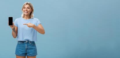 Guarda a Questo divertente foto. intrattenuto attraente contento donna con giusto capelli nel casuale maglietta e denim pantaloncini mostrando smartphone a telecamera puntamento a dispositivo schermo sorridente a grandi linee al di sopra di blu parete foto