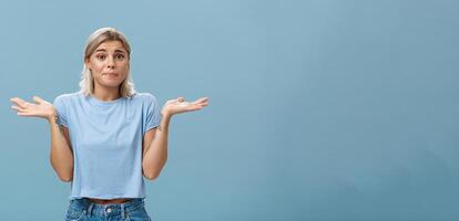 no idea Come Aiuto spiacente. ritratto di insicuro cupo fidanzata sensazione rimpiangere alzando le spalle con mani vicino le spalle accigliato e inseguendo labbra essere incerto e ignaro al di sopra di blu sfondo foto