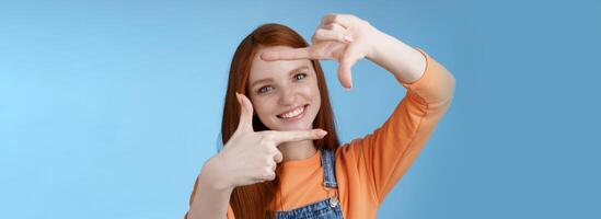 gioioso attraente sincero testa Rossa giovane ragazza ricerca ispirazione trova Perfetto angolo prendere bene tiro rendere mano montatura Guarda attraverso Contentissimo divertito sorridente a grandi linee bianca denti, blu sfondo foto