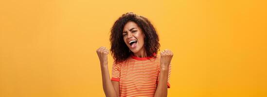 allegro Contentissimo e entusiasta africano americano donna con afro acconciatura stringendo sollevato pugni urla sì a partire dal trionfo e gioia di successo in piedi soddisfatto di vittoria al di sopra di arancia parete foto