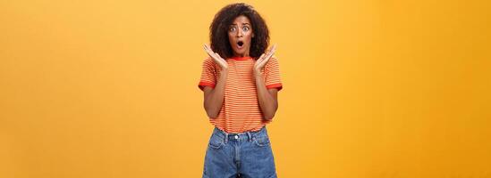 Oh no che cosa accaduto. preoccupato africano americano sorella con Riccio capelli nel di moda a strisce maglietta e denim pantaloncini ansimando lancio mascella, guardare ha riguardato esprimendo empatia e shock al di sopra di arancia parete foto