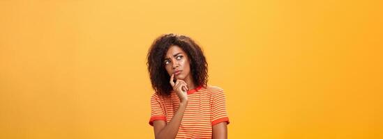 donna considerando per prendere parte nel evento. inteligente focalizzata e creativo bella vista africano ragazza con Riccio capelli Tenere dito su labbro guardando ha riguardato sinistra, pensiero fabbricazione decisione al di sopra di arancia parete foto