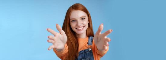 venire in braccia. affascinante sincero contento genere testa Rossa ragazza bambino seduta allungare mani telecamera voglio hold catturare sorridente amichevole chiede passaggio oggetto, in piedi blu sfondo raggiungere amico dare coccole foto