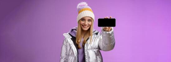 fiducioso amichevole bella vista biondo ragazza nel all'aperto argento luccicante giacca cappello hold smartphone orizzontale mostrando mobile Telefono Schermo assertivo Sorridi raccomandare uso app, viola sfondo foto