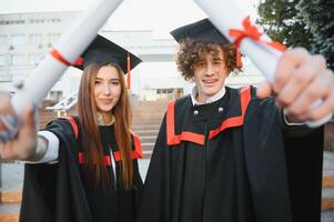 ritratto di contento laureati. Due amici nel la laurea caps e abiti in piedi al di fuori Università edificio con altro studenti nel sfondo, Tenere diploma pergamene, e sorridente foto