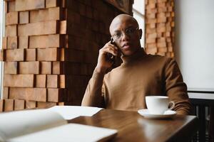 africano uomo d'affari parlando su Telefono seduta a bar tavolo, occupato imprenditore Lavorando distante nel caffè Casa con il computer portatile documenti A proposito di su mobile, nero uomo fabbricazione chiamata avendo pranzo nel caffetteria. foto