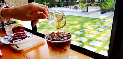 vicino su mano di persone scrosciante corto di buio caffè espresso caffè o tè nel bicchiere di ghiaccio bibita con dolce rosso torta e verde erba sfondo. rinfresco potabile concetto. foto