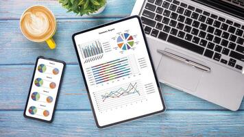 un' dinamico spazio di lavoro in mostra un' tavoletta e smartphone con statistico grafici, a fianco un' computer portatile, caffè, e penna su un' blu di legno tavolo. foto