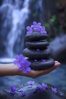 ai generato un' mano Tenere un' pila di zen pietre, circondato di viola fiori, con un' sfocato cascata nel il sfondo, la creazione di un' tranquillo e sereno scena. foto