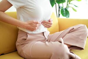 bellissimo asiatico donna con addominale dolore nel il mezzo sopra il ombelico. lei si siede su il divano a casa. concetto di trattamento di pancreas, stomaco, cistifellea o esofago nel donne. Salute cura foto