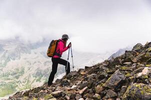 donna viaggiatore va per il superiore di il montagna. un' ragazza con il trekking poli e un' zaino fermato per ammirare il nebbia si avvicina sua nel il montagne foto