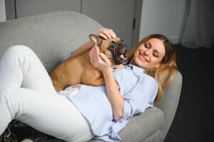 giovane bella donna nel casuale Abiti abbracciare sua cane seduta su il divano nel il vivente camera di sua accogliente nazione Casa. animale comunicazione concetto foto