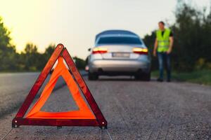 irriconoscibile triste autista nel riflessivo veste. maschio autista in piedi vicino rotto auto con Aperto su cappuccio. rosso triangolo per avvisare altro strada utenti di auto abbattersi o motore fallimento fermare a campagna autostrada. emergenza traffico situazione foto