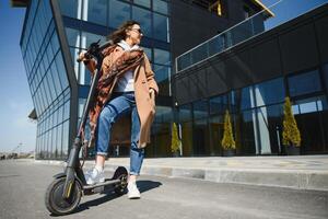 giovane bellissimo donna nel un' giacca sorrisi e cavalcate un elettrico scooter per opera lungo ufficio edifici foto