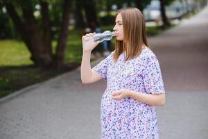 incinta donna sta nel il parco con un' bottiglia di acqua. copia spazio foto