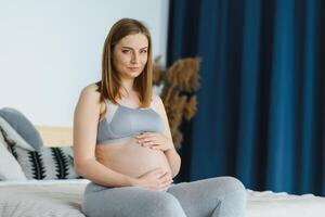 attraente incinta donna è seduta nel letto e Tenere sua gonfiarsi. ultimo mesi di gravidanza. incinta giovane ragazza. assistenza sanitaria concetto. salutare cibo concetti. rilassante a casa. cura di incinta. foto