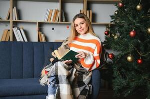 attraente giovane signora con buio capelli lettura interessante libro mentre seduta su grigio divano. sfocatura sfondo di bellissimo Natale albero. accogliente atmosfera. foto