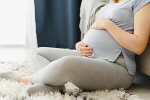 incinta donna carezzevole sua pancia a casa. giovane in attesa bionda sensazione sua bambino spingere, seduta su pavimento, copia spazio. gravidanza, riposo, vita, aspettativa concetto foto