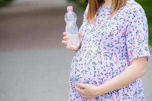 incinta donna sta nel il parco con un' bottiglia di acqua. copia spazio foto