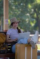 ragazza utilizzando un' il computer portatile mentre in attesa nel un' treno stazione, ragazza su treno stazione con bagaglio Lavorando su il computer portatile computer, il computer portatile nel uso, si siede con un' valigia foto