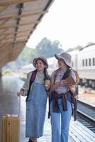 Due giovane asiatico amici ragazze con zaini a ferrovia stazione in attesa per treno, Due bellissimo donne a piedi lungo piattaforma a treno stazione foto