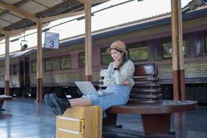 ragazza utilizzando un' il computer portatile mentre in attesa nel un' treno stazione, ragazza su treno stazione con bagaglio Lavorando su il computer portatile computer, il computer portatile nel uso, si siede con un' valigia foto