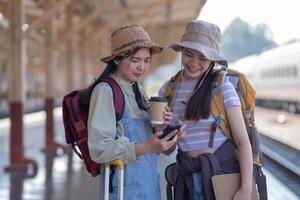 Due giovane asiatico amici ragazze con zaini a ferrovia stazione in attesa per treno, Due bellissimo donne a piedi lungo piattaforma a treno stazione foto