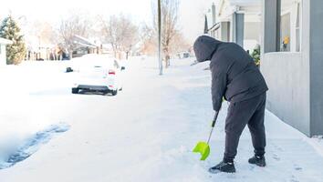 un' uomo nel un' caldo nero giacca e i pantaloni spalare neve nel davanti di loro casa per preparare il strada per convenienza. foto