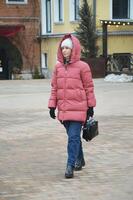 un' giovane elegante caucasico donna nel un' giù giacca, a maglia cappello e jeans passeggiate lungo un' città strada nel inverno. foto