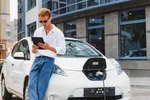 ritratto di giovane bello uomo nel casuale indossare, in piedi a il ricarica stazione. eco elettrico auto concetto foto