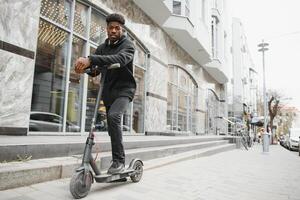 afro americano tipo cavalcate un elettrico scooter contro il sfondo di un' parete, un' alunno usi eco trasporto foto