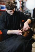 lato Visualizza di grave uomo con elegante moderno taglio di capelli guardare inoltrare nel barbiere negozio. mano di barbiere conservazione dritto rasoio e taglio di moda strisce su testa di cliente. foto