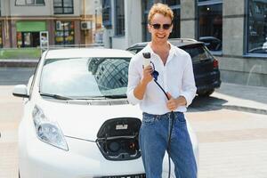 uomo Tenere energia ricarica cavo per elettrico auto nel all'aperto auto parco. e lui S andando per Collegare il auto per il ricarica stazione nel il parcheggio lotto vicino il shopping centro foto