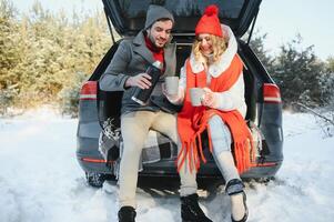 coppia nel amore seduta nel auto tronco potabile caldo tè nel nevoso inverno foresta e chattare. persone rilassante all'aperto durante strada viaggio. san valentino giorno foto
