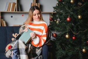 attraente giovane signora con buio capelli lettura interessante libro mentre seduta su grigio divano. sfocatura sfondo di bellissimo Natale albero. accogliente atmosfera. foto