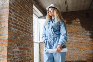 attraente femmina costruzione lavoratore nel elmetto protettivo. fiducioso giovane specialista nel scacchi blu camicia nel jeans in piedi nel vuoto camera. interno design e rinnovamento servizio. foto