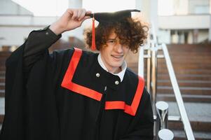 la laurea a partire dal Università. giovane sorridente ragazzo Università diplomato nel tradizionale bonet e mantello in piedi e Tenere diploma nel mano al di sopra di Università edificio sfondo foto