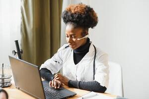 sorridente africano americano femmina medico gp indossa bianca medico cappotto utilizzando il computer portatile computer a posto di lavoro dà a distanza in linea consultazione, Lavorando su pc, consulenza paziente nel Internet telemedicina Chiacchierare foto