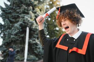 contento sorridente Università diplomato nel mantello Tenere diploma nel sollevato mano e esprimendo felicità al di sopra di Università edificio a sfondo. riuscito laurea a partire dal Università o Università foto