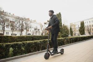 afro americano tipo cavalcate un elettrico scooter contro il sfondo di un' parete, un' alunno usi eco trasporto foto