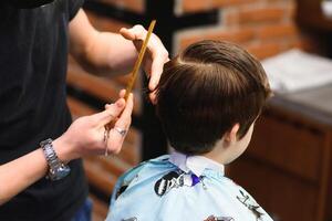 un' poco carino ragazzo si siede nel un' parrucchiere a il dello stilista, un' scolaro è ottenere capelli tagliare nel un' bellezza salone, un' bambino a un' dal barbiere, un' corto Uomini taglio di capelli foto