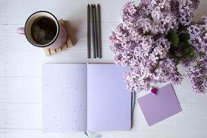 bloc notes con matita, lilla nel vaso e un' tazza di caffè su un' bianca di legno desktop. ispirazione posto di lavoro. foto